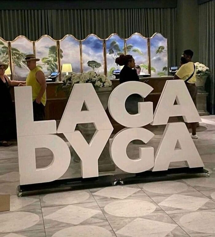 Large stacked letters spelling lagadyga in an awkward photo of a lobby with people in the background.