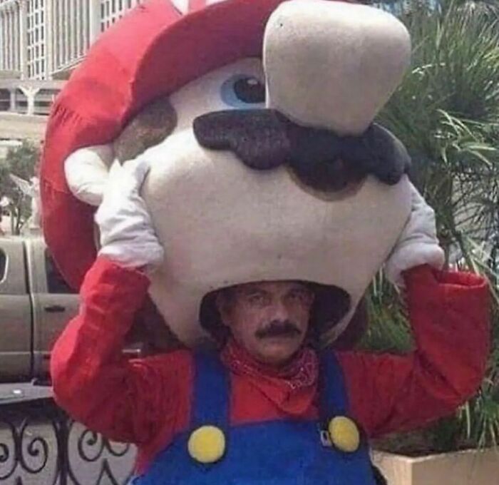 Man awkwardly wearing oversized Mario costume head, struggling to keep it on during outdoor event.
