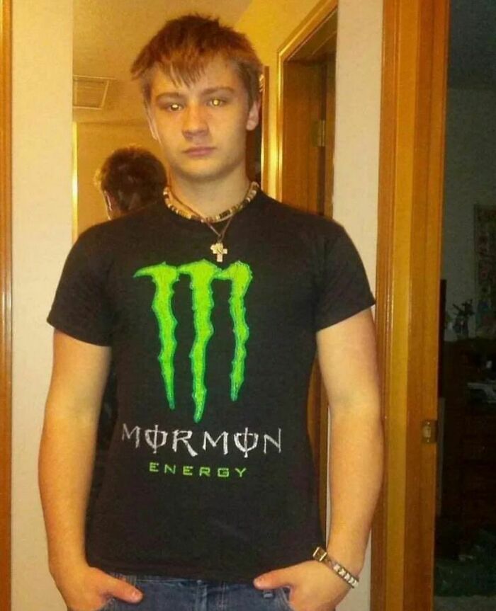 Young man wearing a black shirt with a parody logo standing awkwardly in a home hallway, awkward photos concept.