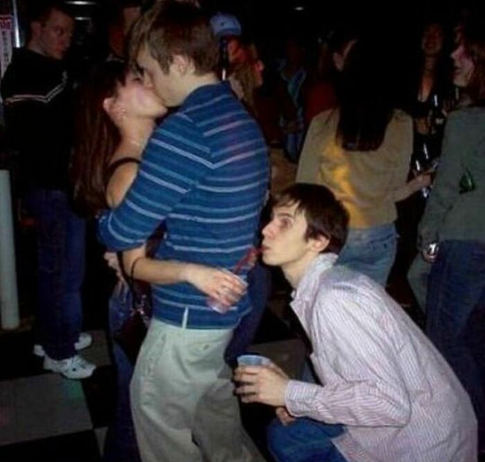 Young man awkwardly sipping drink behind a couple kissing at a crowded party, capturing an awkward moment.