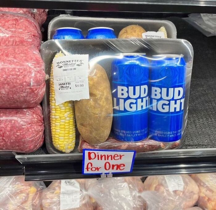 Packaged dinner for one with corn, potato, two cans of beer, and meat awkwardly displayed in grocery store.