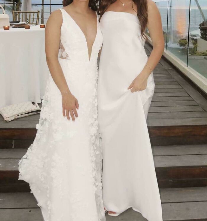 Two brides in white gowns standing side by side on a wooden deck at a wedding ceremony.