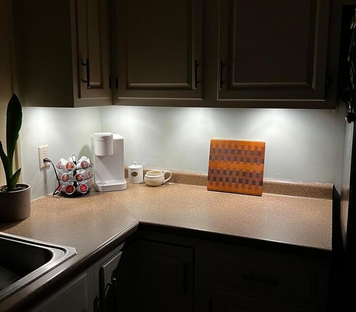 Under-cabinet kitchen lighting illuminating countertop with coffee maker, pod holder, cup, and cutting board, showcasing useful random things.