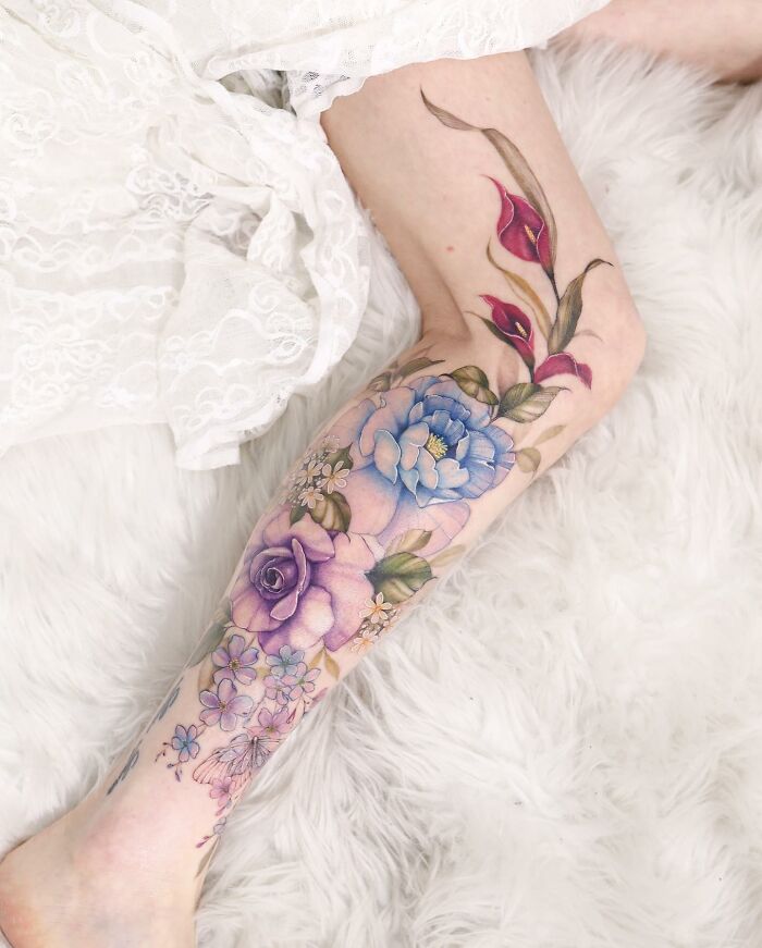 Watercolor-like tattoo design on a leg, featuring vibrant flowers and leaves on a soft, textured background.