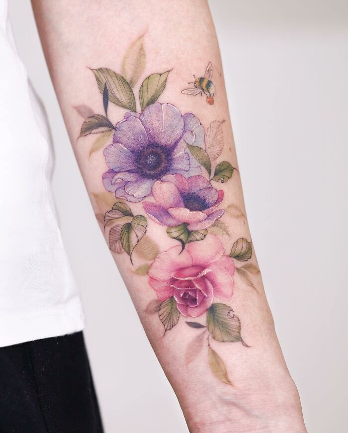 Watercolor-style tattoo design on an arm featuring purple and pink flowers with a bee.