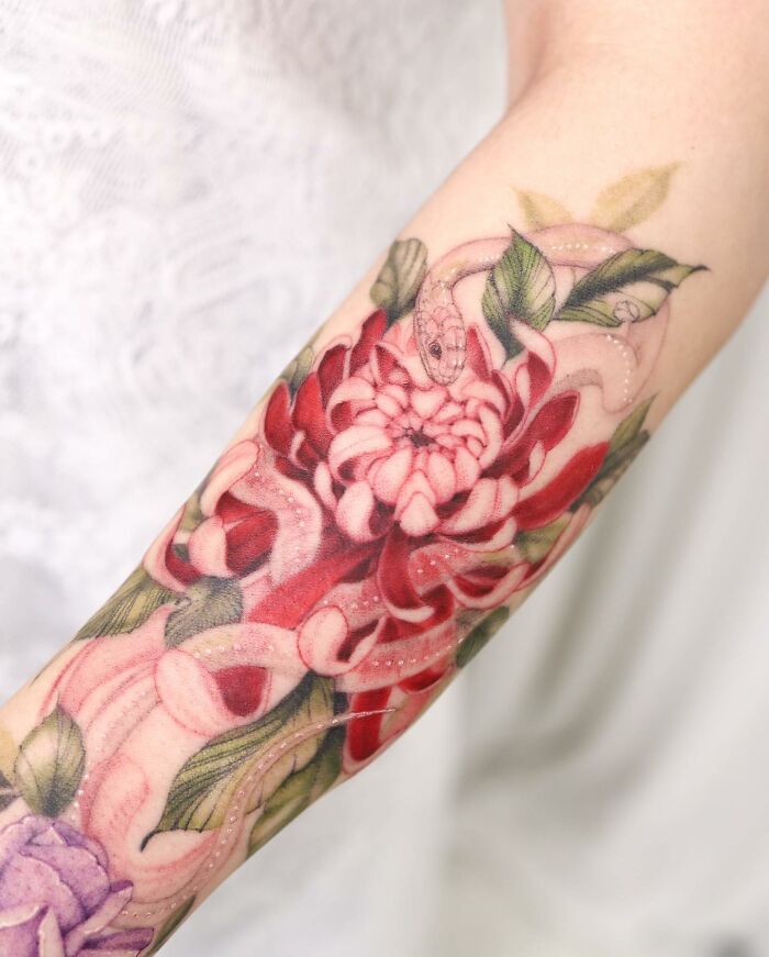 Watercolor tattoo design on forearm featuring a pink flower and swirling green leaves.