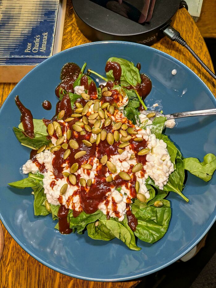 A strange dish with spinach, cottage cheese, barbecue sauce, and pumpkin seeds on a blue plate.