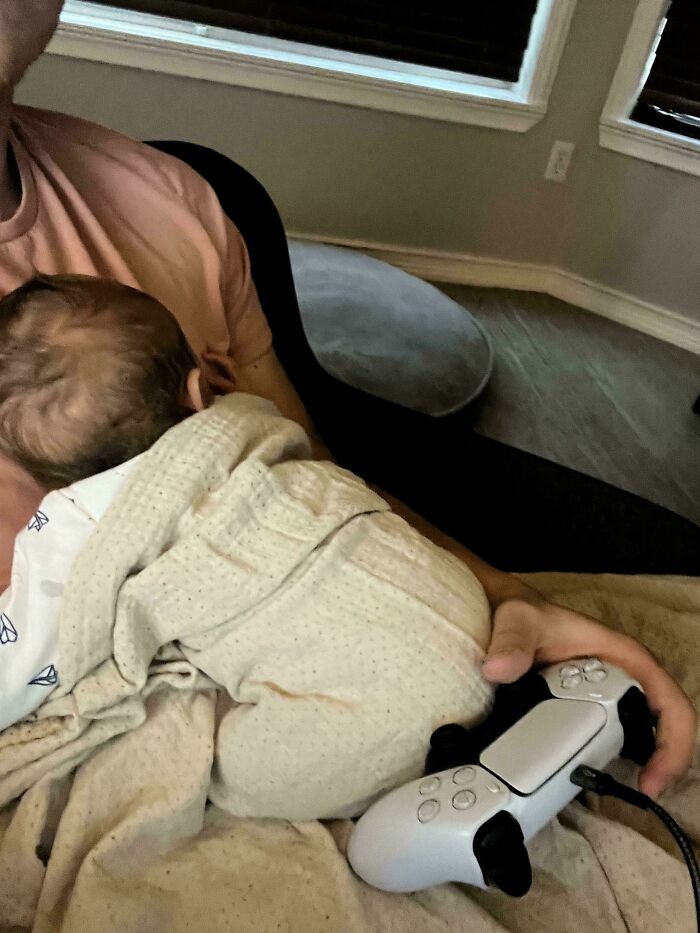 Dad holding a baby on his lap while gaming with a controller, showing a wholesome and goofy dad moment.