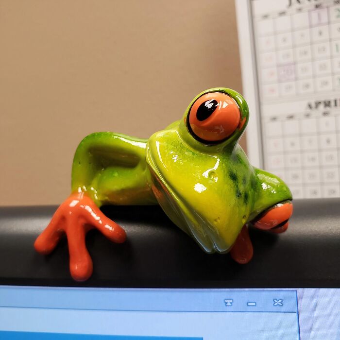 A colorful frog figurine perched on a computer monitor, brightening work supplies in an office setting.