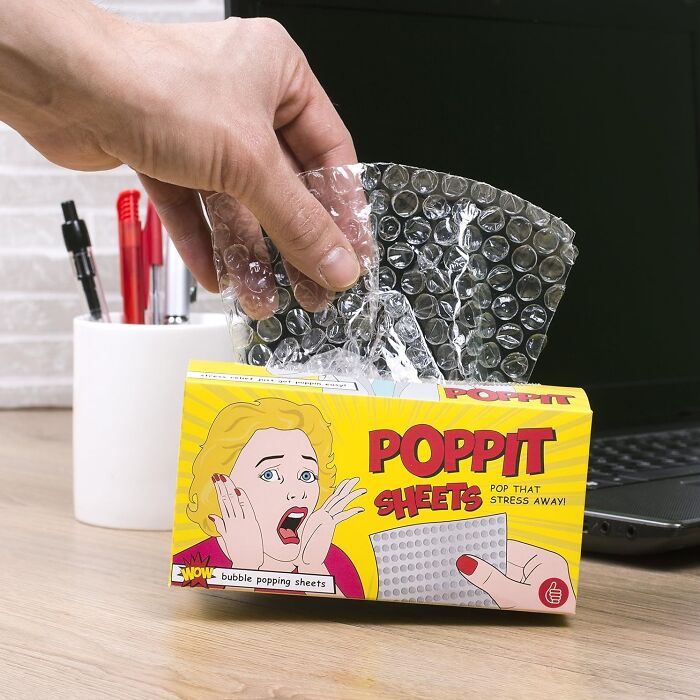 Hand holding Poppit bubble wrap, a stress-relieving work supply, near a laptop.
