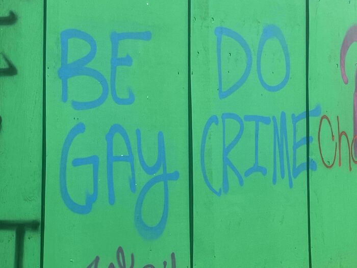 Sign fail with graffiti on a green wall reading "BE GAY DO CRIME" in blue letters.