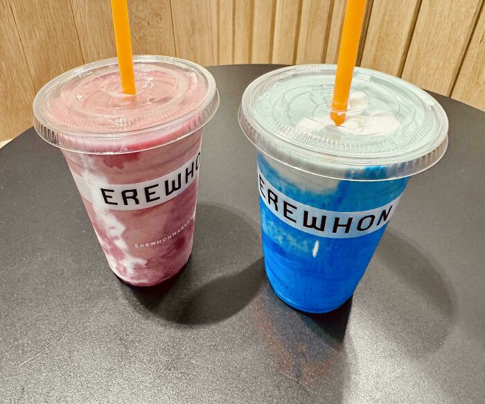 Two Erewhon smoothies, one pink and one blue, with orange straws on a dark table.