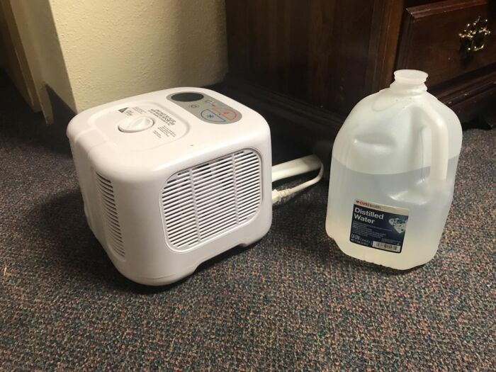 White home humidifier and gallon of distilled water placed on a carpeted floor near wooden furniture for indoor comfort.