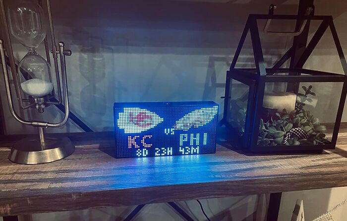 Digital display of KC vs PHI game scores on a shelf with hourglass and plant, part of a collection of random things.