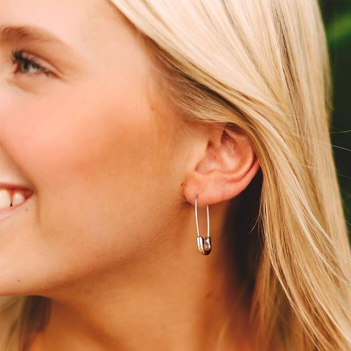Close-up of a person wearing safety pin earrings, smiling and showing blonde hair.