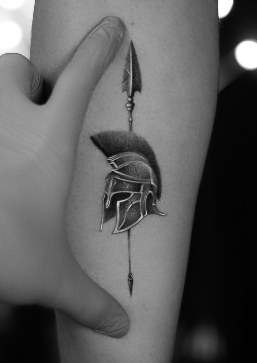 Single needle tattoo of Spartan helmet with spear on forearm.