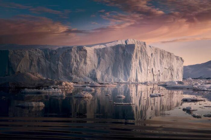 Massive iceberg reflecting on calm water under a colorful sky, showcasing breathtaking landscape photography from 2024 awards.