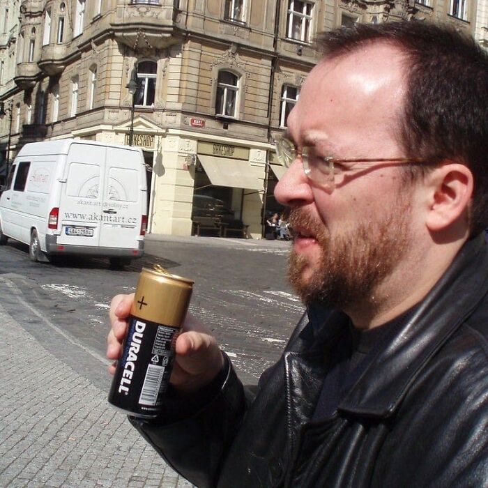 Man in black jacket holding oversized Duracell battery in city street, fitting cursed pics and memes theme from Instagram page.