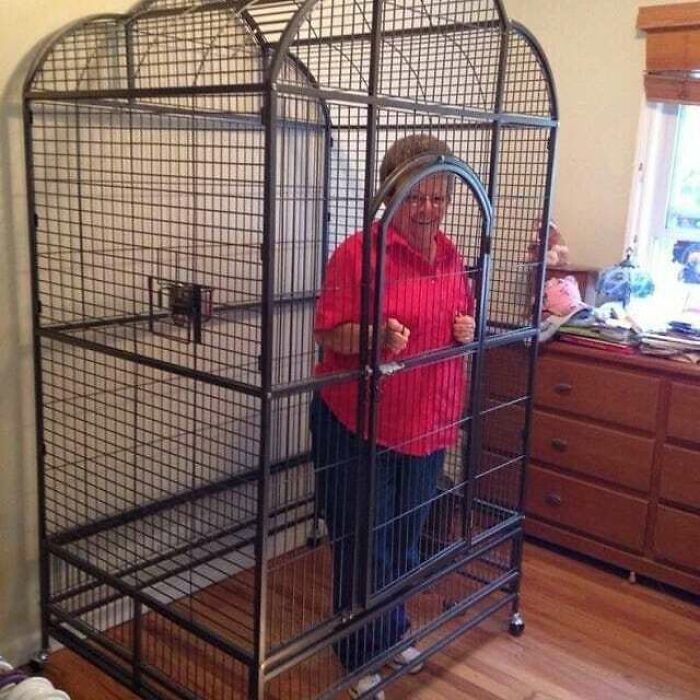 Elderly woman standing inside a large empty metal cage in a room, a cursed pic from Instagram meme page.