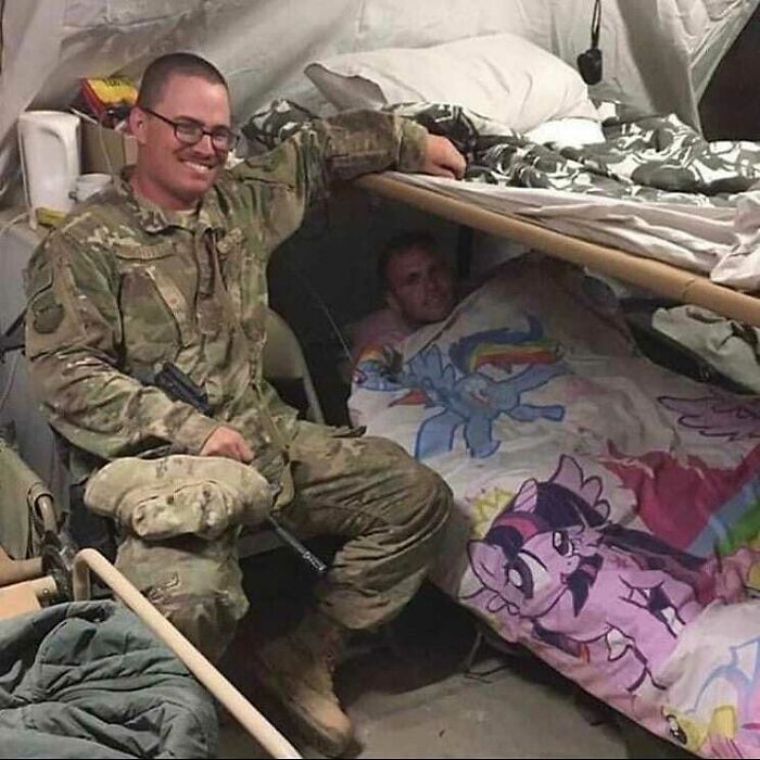 A soldier smiling next to a bunk bed with a person under a colorful My Little Pony blanket, cursed pics and memes.