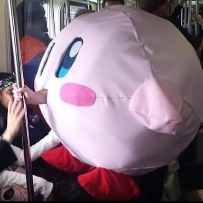 Person wearing a large pink costume with cartoon eyes and blush, sitting on a crowded subway, sharing cursed pics and memes.