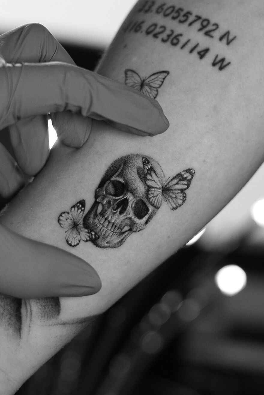 Single needle tattoo of a skull with butterflies on an arm, artist wearing gloves, showcasing intricate details.