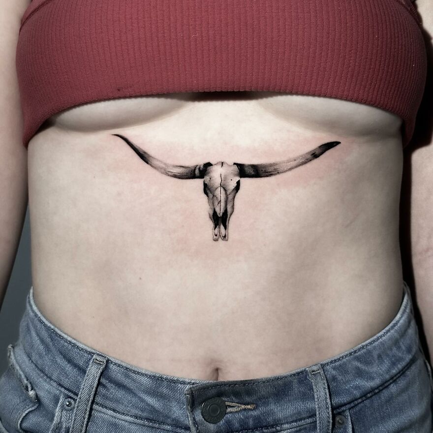 Single needle tattoo of a bull skull on a person's stomach, wearing a red crop top and blue jeans.