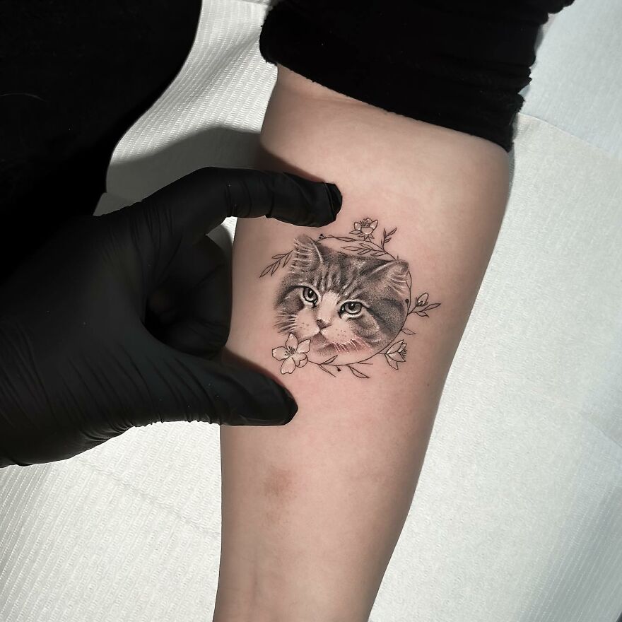 Single needle tattoo of a cat with floral design on forearm, artist wearing black gloves.