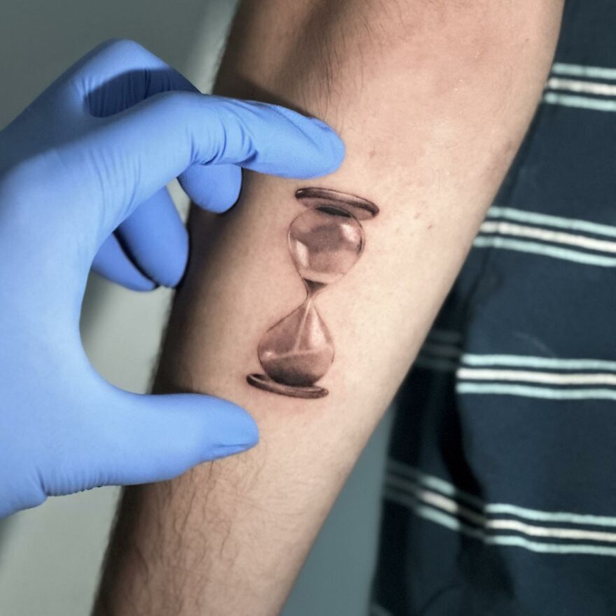 Single needle tattoo of an hourglass on an arm, detailed in black ink, with a gloved hand adjusting the skin.