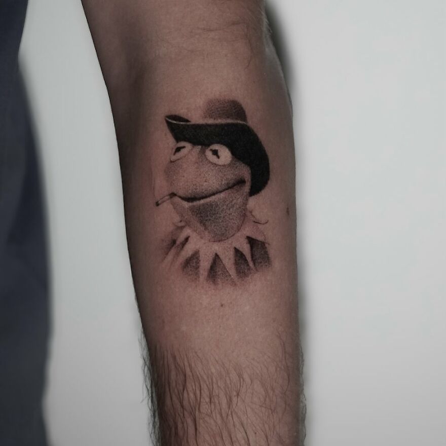 Single needle tattoo of Kermit the frog character wearing a hat on a person's arm.