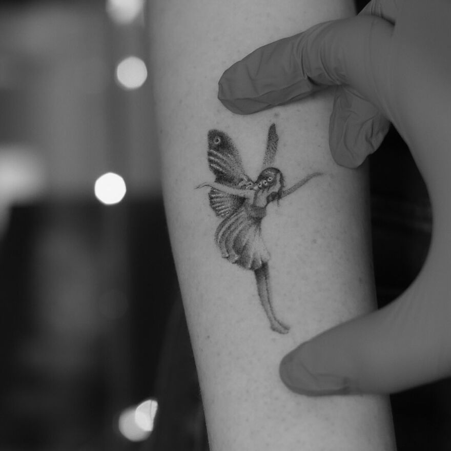 A single-needle tattoo of a fairy on an arm, with a gloved hand nearby.