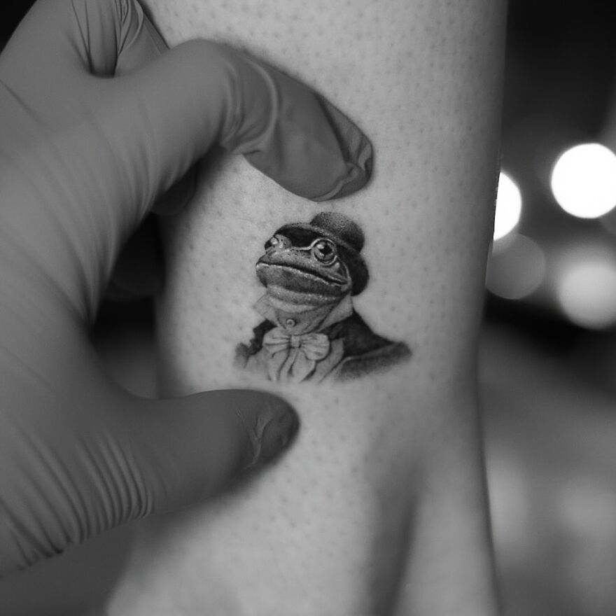 Single needle tattoo of a frog in a suit and top hat, being gently held by a gloved hand.