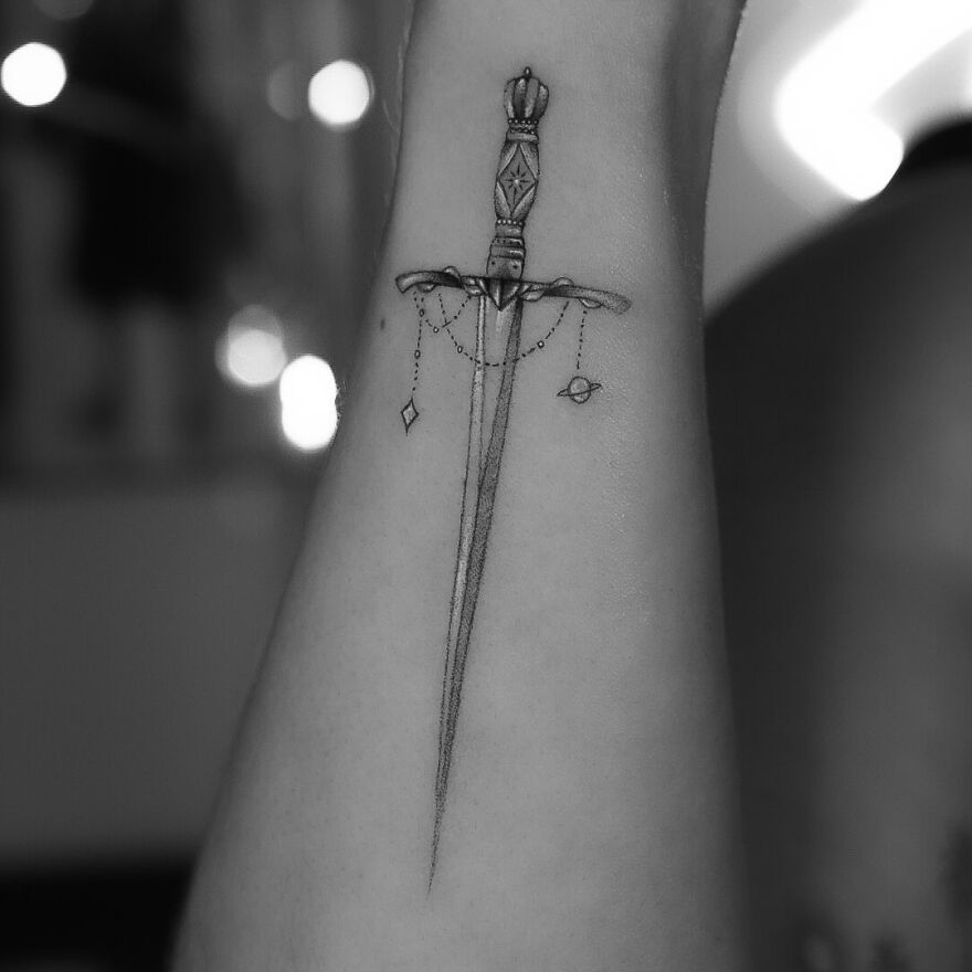Single needle tattoo of an ornate sword with delicate chains on an arm.