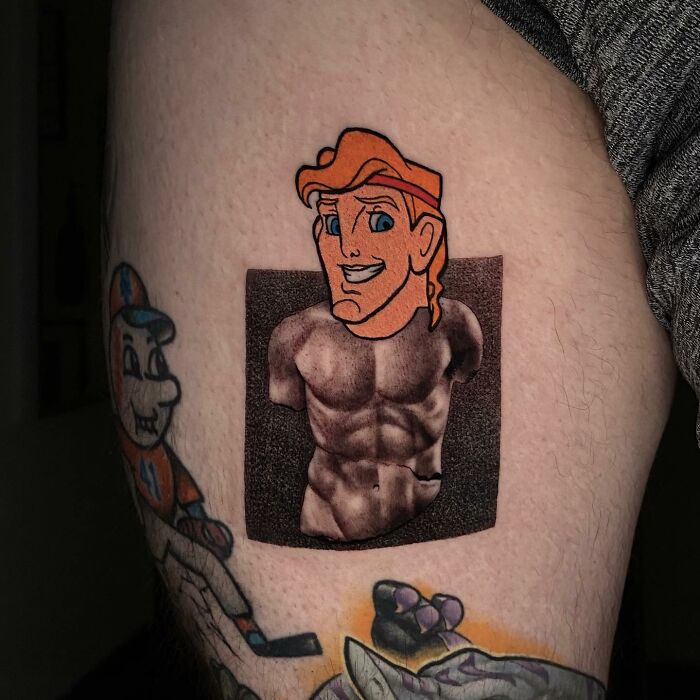 Tattoo merging cartoon and classical art styles on a person's upper leg, featuring a character with muscular torso.