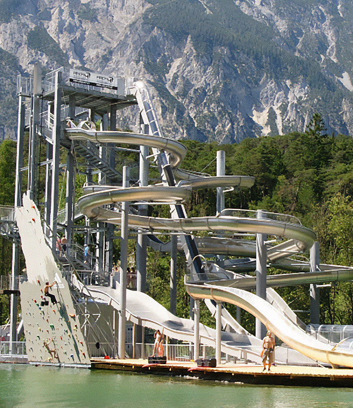 Extreme waterslide with thrill-seeking diver jumping down, surrounded by mountainous landscape and dense forest backdrop.