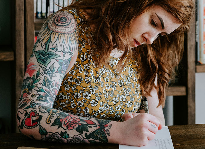 Woman with dark tattoo sleeves writing on a piece of paper.