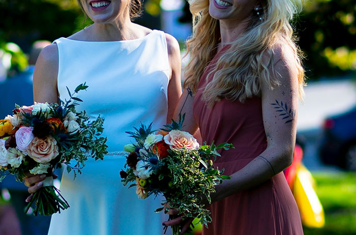 Bride and MOH holding bouquets, MOH has visible dark tattoos on her arm, both in elegant dresses.