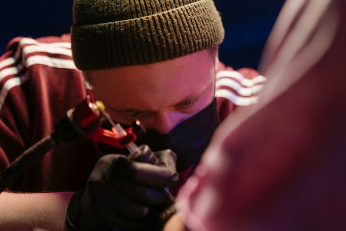 Tattoo artist focused on creating a detailed design, wearing a beanie and mask.