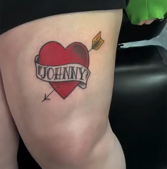 Tattoo of a heart with an arrow, featuring "JOHNNY" on a ribbon, showcasing an uncomfortable creation.