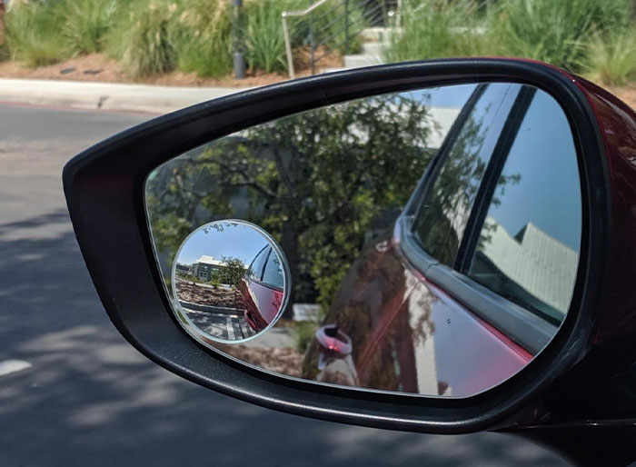 Car mirror with blind spot mirror attachment reflecting street view.