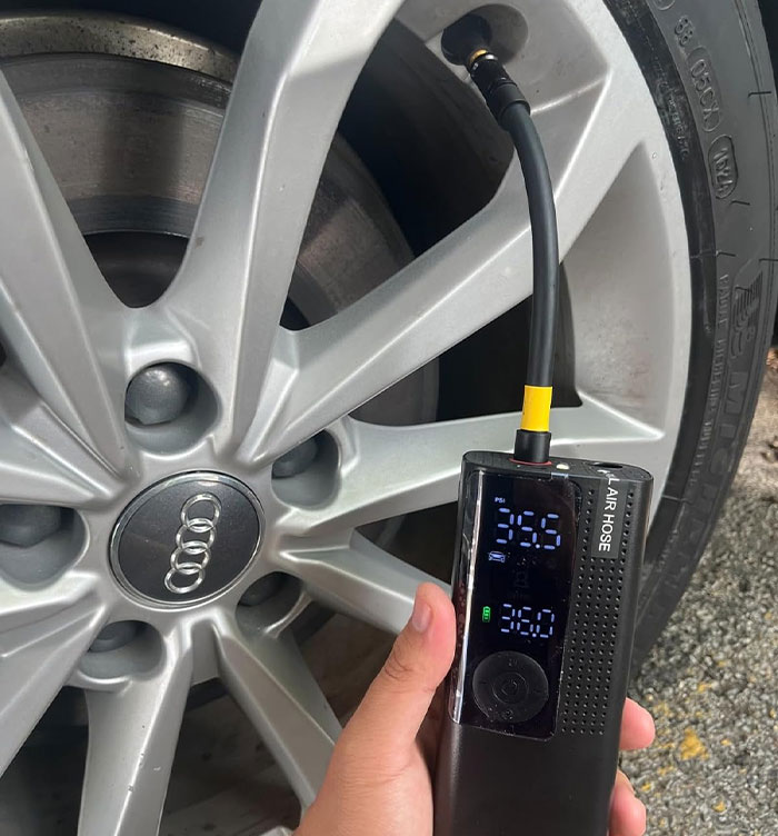 Portable car tire inflator held by person, displaying pressure settings, with a background of a car wheel.