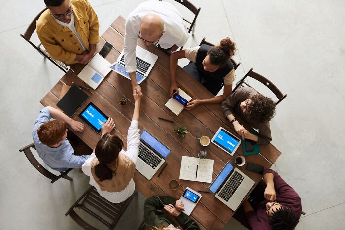 A diverse group of people in their 40s, 50s, and 60s collaborating around a table with laptops and tablets.