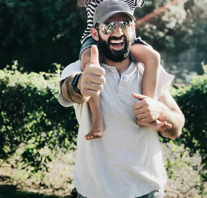 Man wearing sunglasses and cap giving a thumbs up, carrying child on shoulders, epitomizing dad humor outdoors.