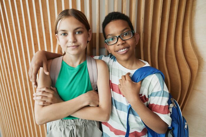 Two smiling Gen Z children with backpacks standing close together against a wooden wall background