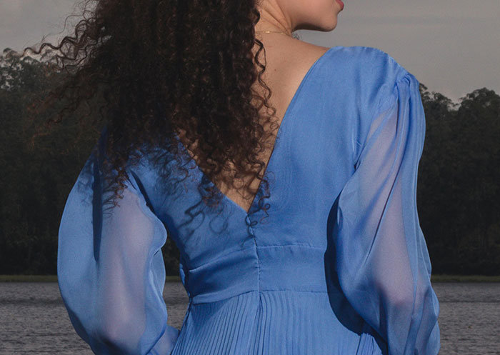 Woman in a blue dress by a lake, with curly hair visible from the back, under a cloudy sky.