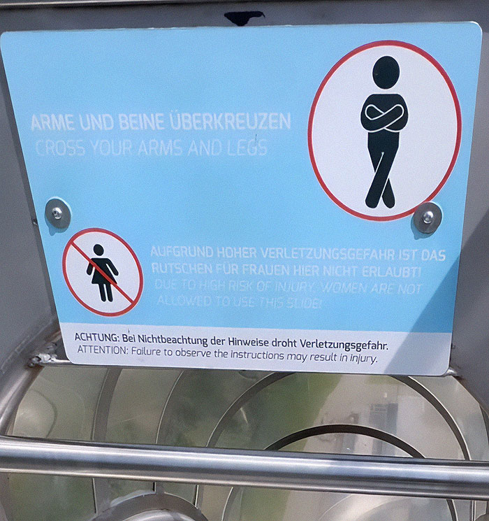 Warning sign at extreme waterslide showing women are banned due to high injury risk and instructions to cross arms and legs.