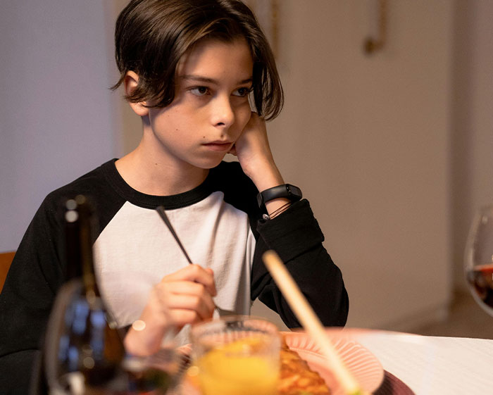 Young boy sitting at a dining table with food and drinks, appearing thoughtful amid strict house rules guests find grimacing.