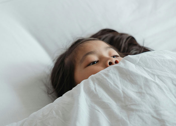 Young girl hiding under white bedding, conveying discomfort and tension related to strict house rules and guests.