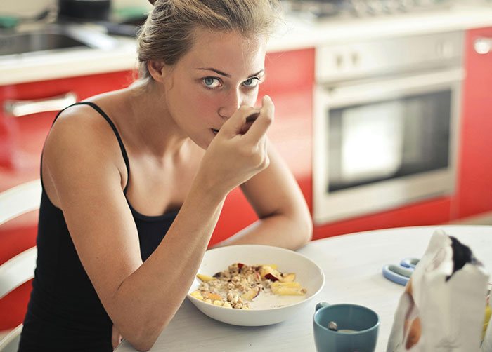Young woman eating breakfast cereal at kitchen table, reflecting household rules that left guests grimacing.