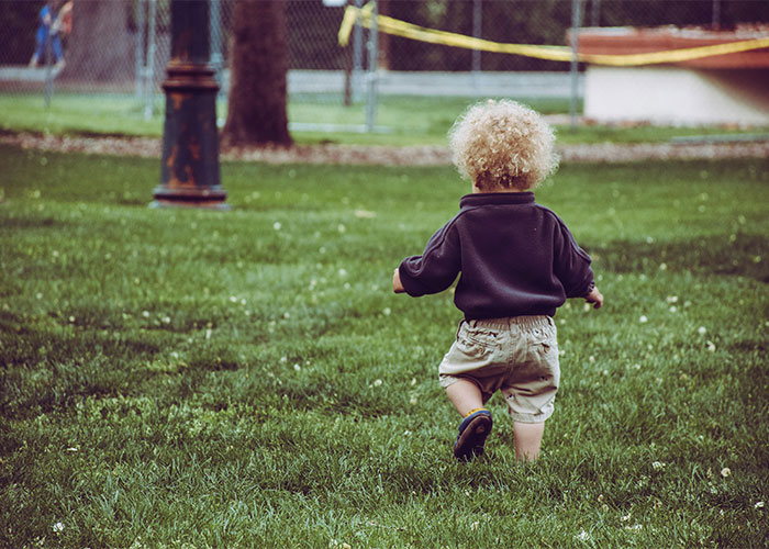 Toddler with curly hair walking on grass in a park, illustrating everyday things most people don’t realize are deadly risks.